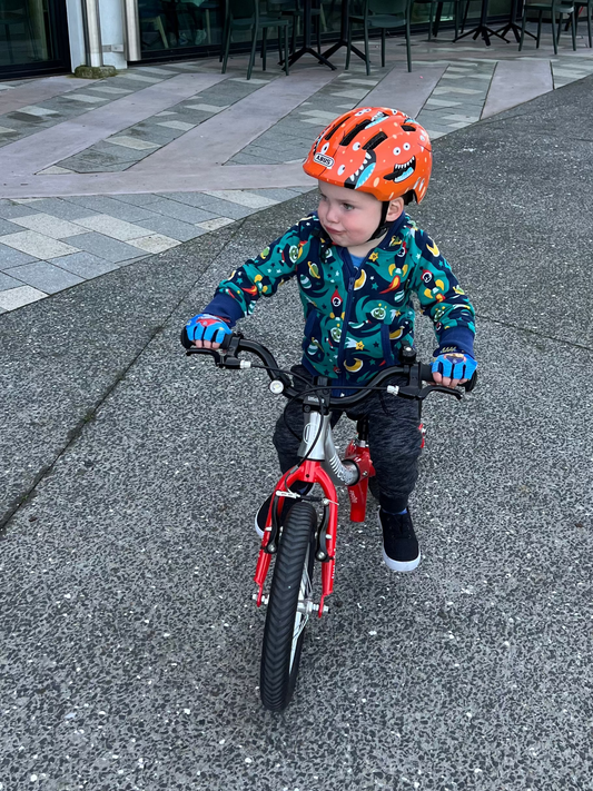 A young child riding a small red balance bike on an outdoor paved path in front of a modern glass-front building. The child is wearing a bright orange helmet, a colorful patterned jacket, and dark pants.