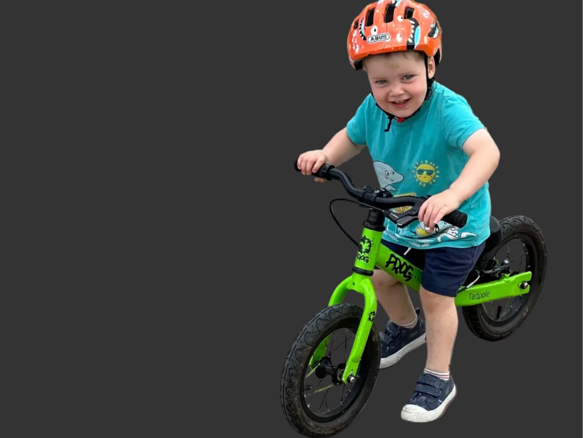 A young child wearing an orange helmet and a turquoise t-shirt rides a bright green balance bike with black tires on a concrete ramp outdoors.