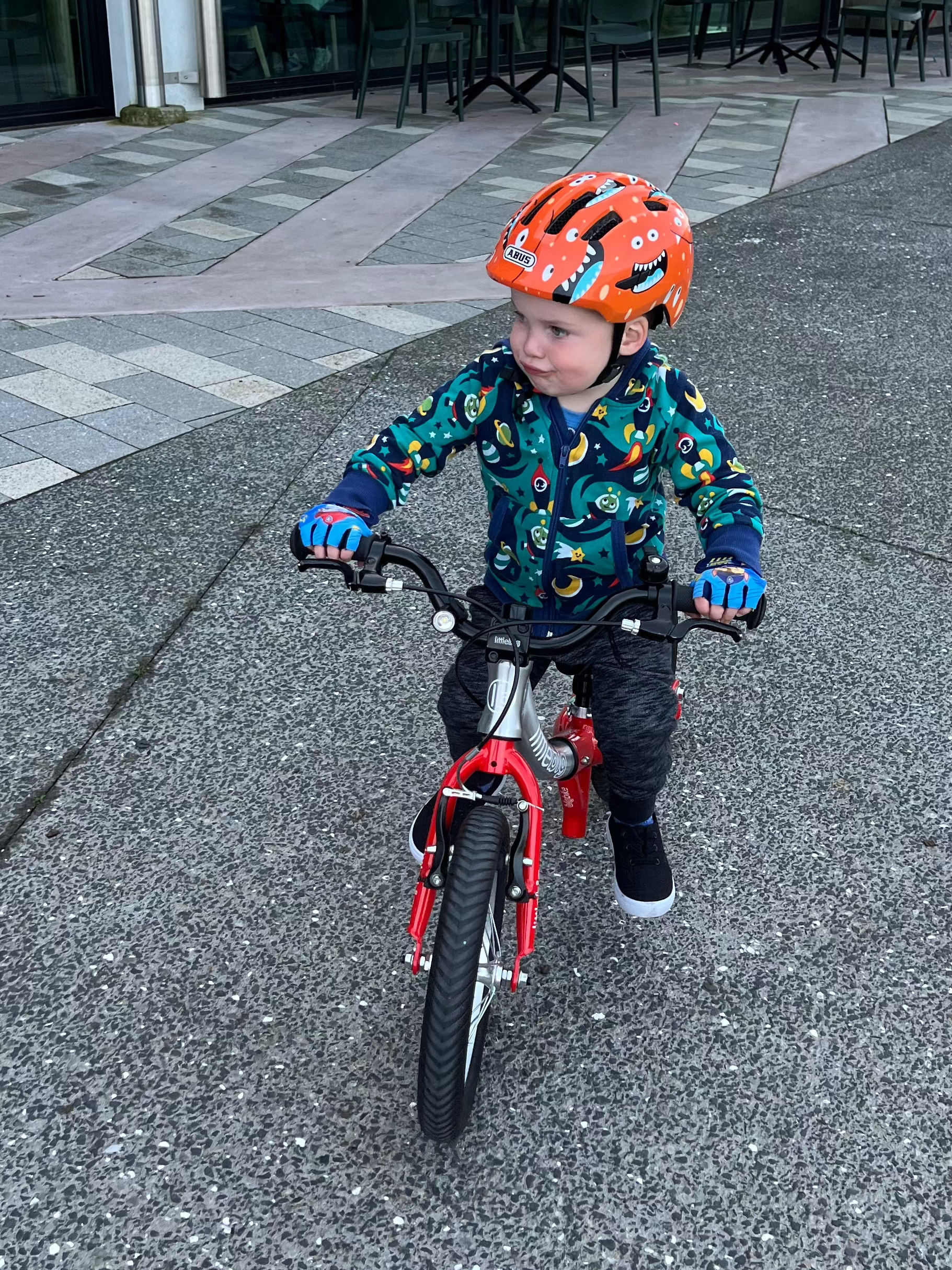 A young child riding a small red balance bike on an outdoor paved path in front of a modern glass-front building. The child is wearing a bright orange helmet, a colorful patterned jacket, and dark pants.