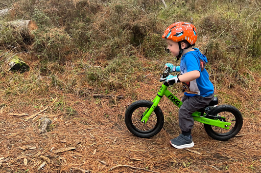 A young child wearing an orange helmet rides a bright green balance bike on a forest trail covered in pine needles and dry grass, with tall trees in the background.