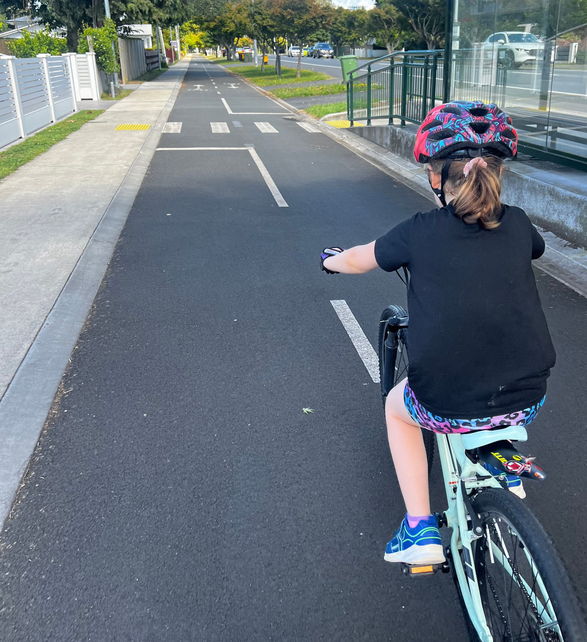 A child wearing a black helmet, black t-shirt, and patterned shorts rides a light blue bicycle on a paved outdoor path under a blue sky.