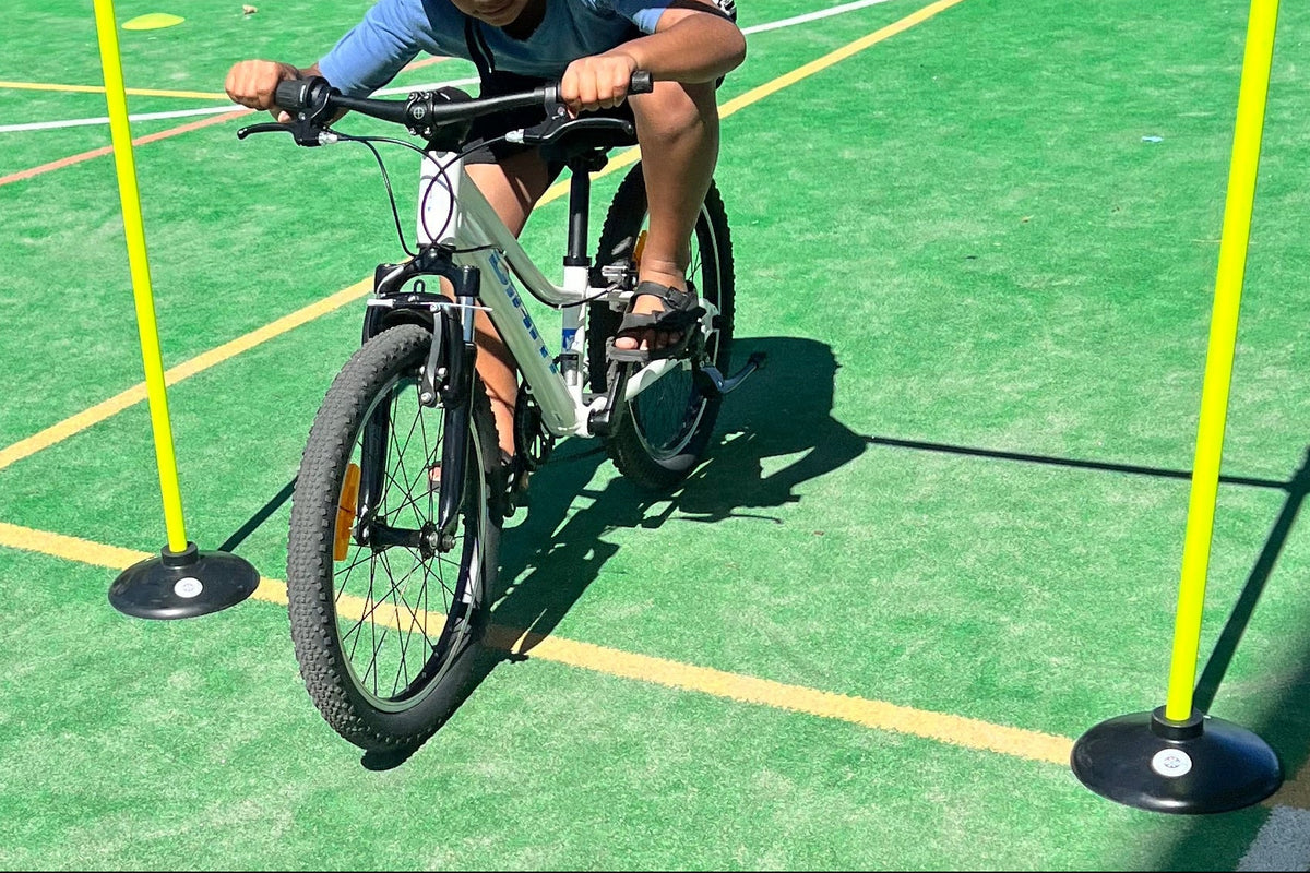 A child wearing a black helmet rides a bicycle through a yellow agility course on a green outdoor court, with other children and trees in the background.