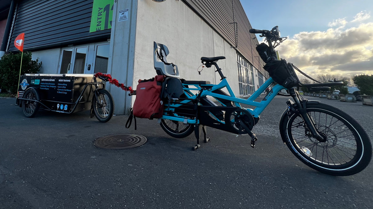 A turquoise electric cargo bike with a red bag and a black utility tricycle attached, parked outdoors in front of a modern dark gray warehouse building under a blue sky.