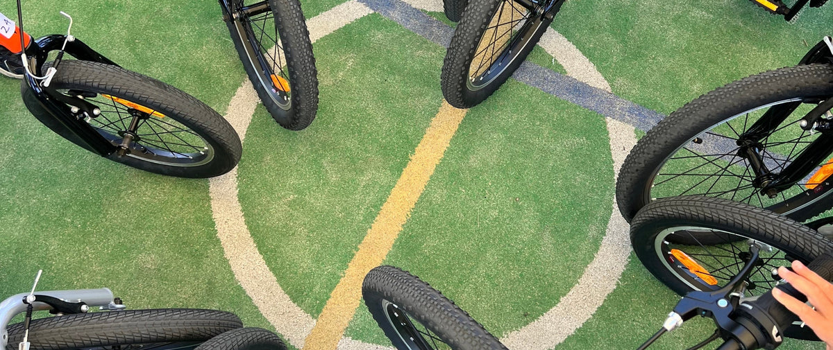 Several children's bicycles are arranged in a circle on a green outdoor court with painted lines, and parts of several people's bodies and hands are visible around the bikes.