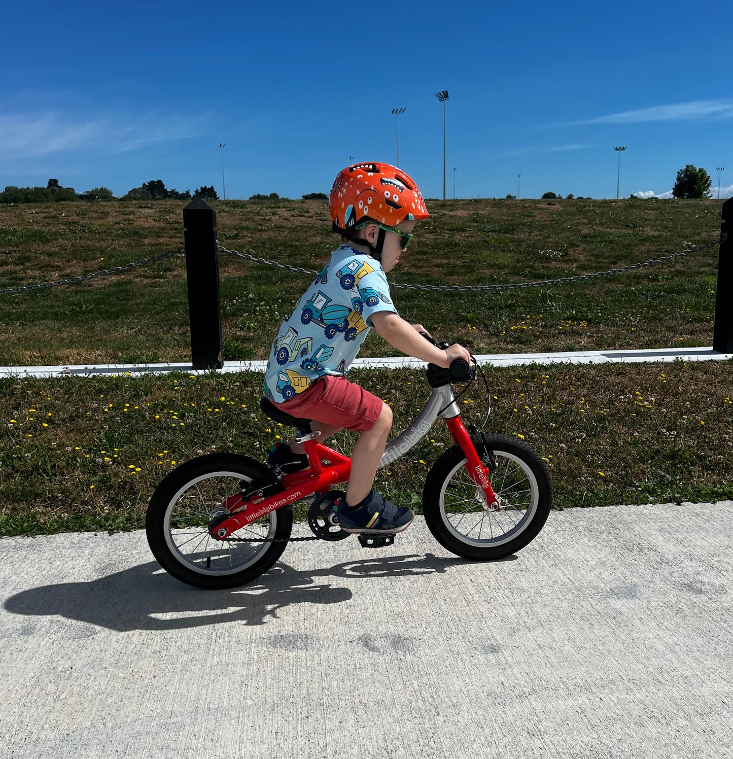 A young child riding a small red balance bike outdoors on a paved path, wearing a patterned shirt, pink shorts, and an orange helmet, with grassy hills and a blue sky in the background.