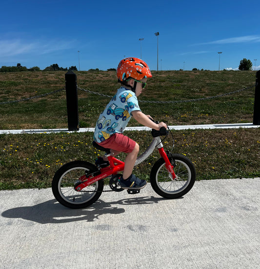 A young child riding a small red balance bike outdoors on a paved path, wearing a patterned shirt, pink shorts, and an orange helmet, with grassy hills and a blue sky in the background.